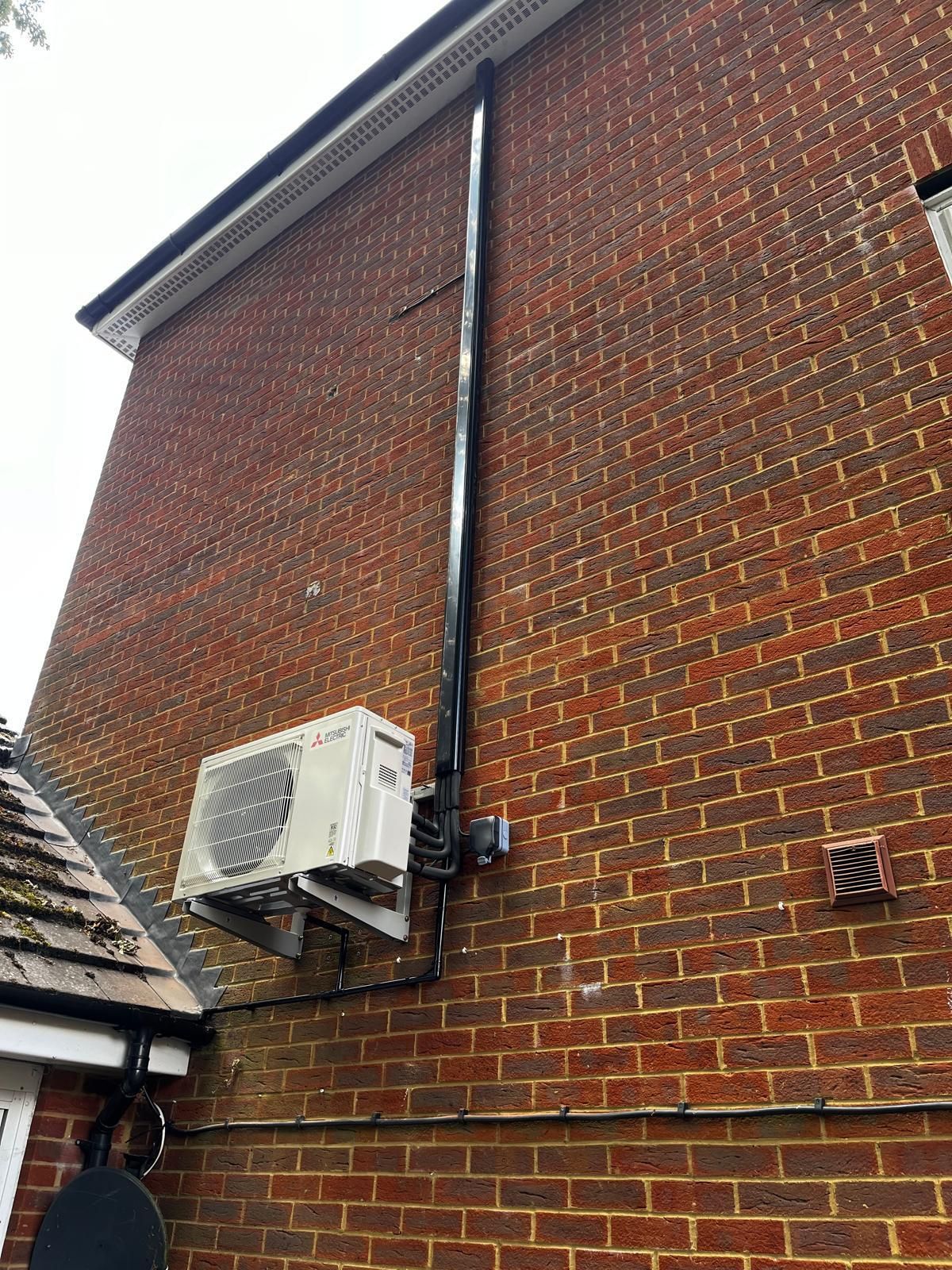 Air conditioning unit mounted on a brick building with black piping running vertically.