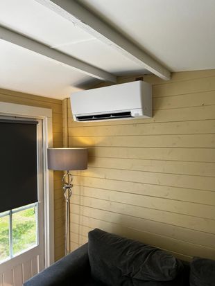 Air conditioner mounted on a yellow-paneled wall, over a sofa and next to a window with a shade.