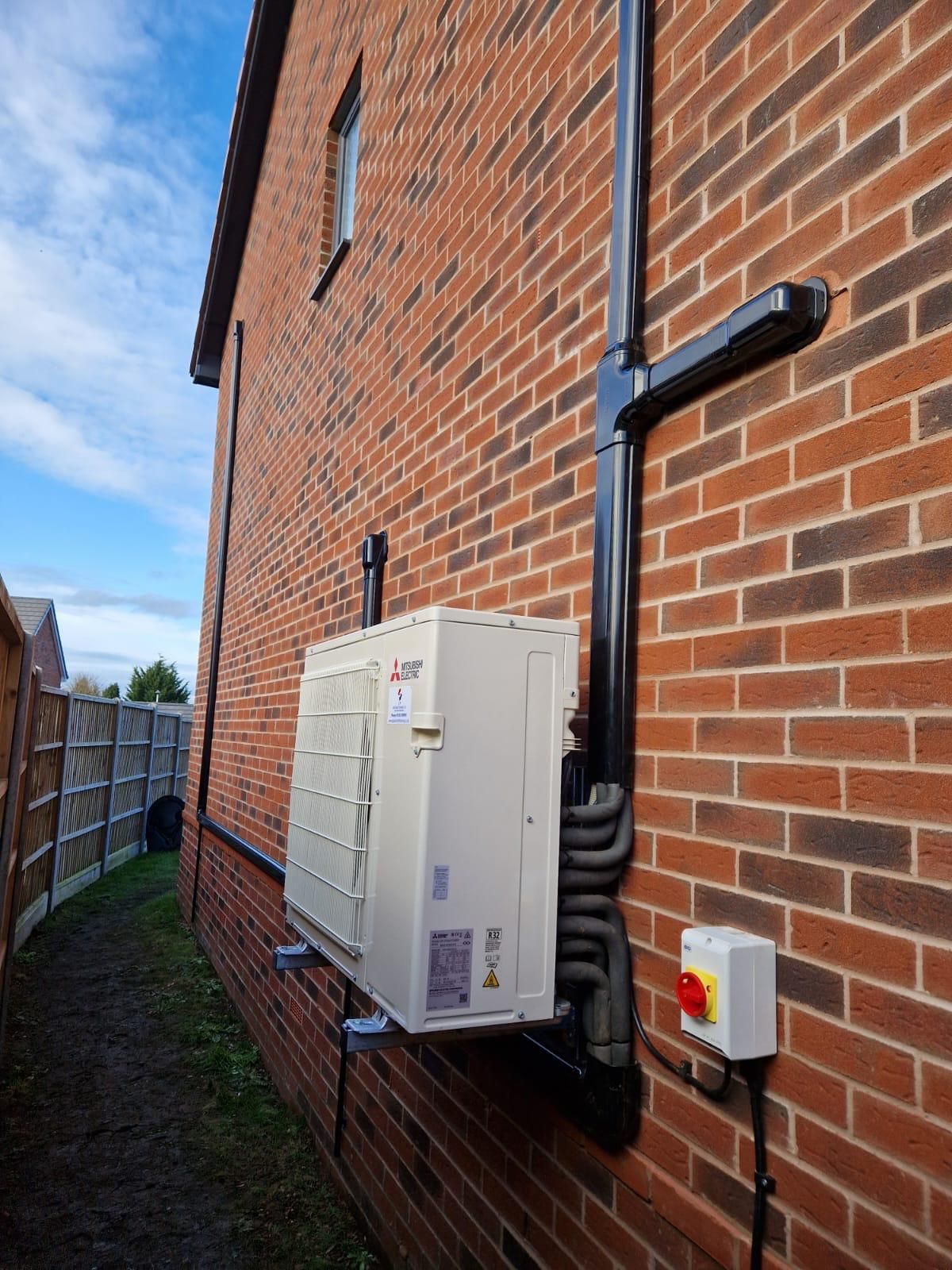 Brick building with an outdoor heat pump unit attached, black piping, and an electrical box.
