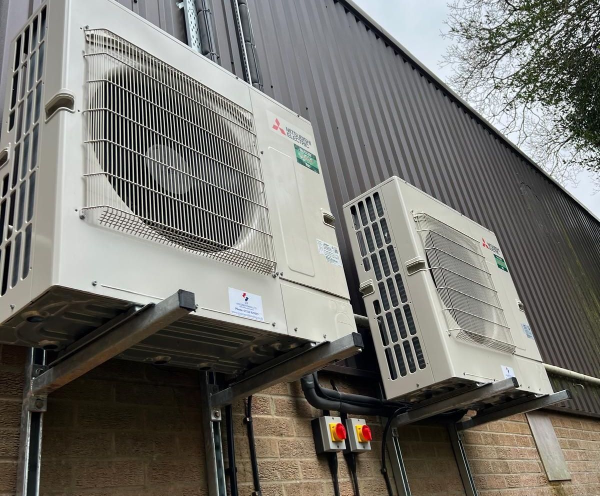 Two white air conditioning units mounted on a brick wall, under a metal roof.