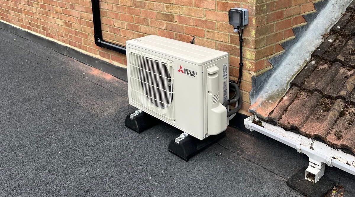 White Mitsubishi air conditioning unit on a flat roof next to a brick wall and a gutter.