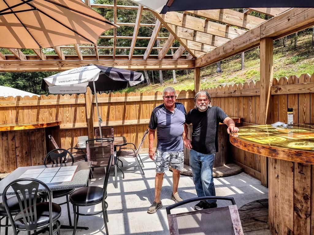 two men are standing in a patio area with tables and chairs under an umbrella that says ' busch ' on it