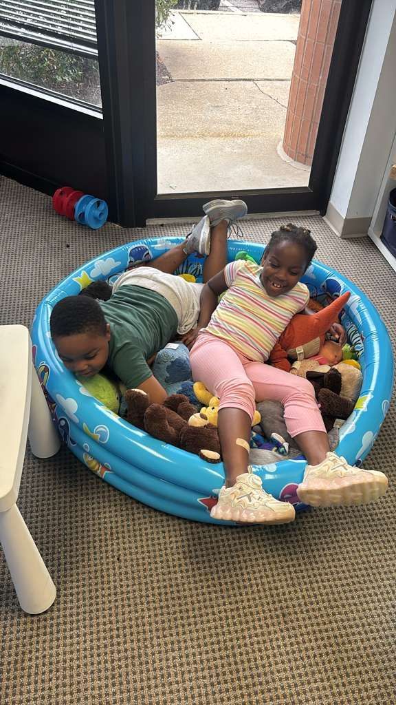Two children in a small blue inflatable pool filled with toys.