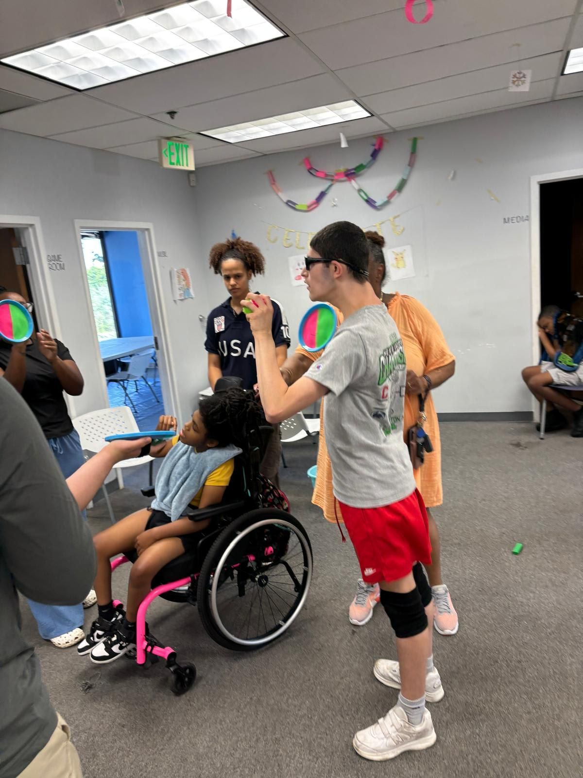 People playing a game in a room. A person in a wheelchair is involved, and others are holding items.