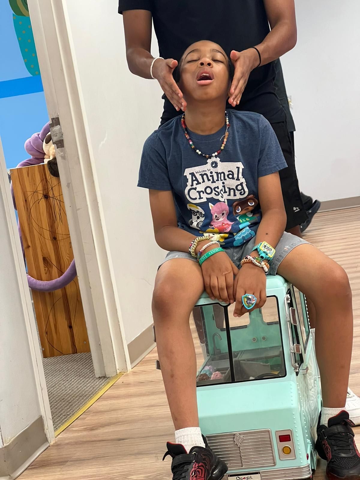 Child on teal toy truck, eyes closed, someone's hands on their head.
