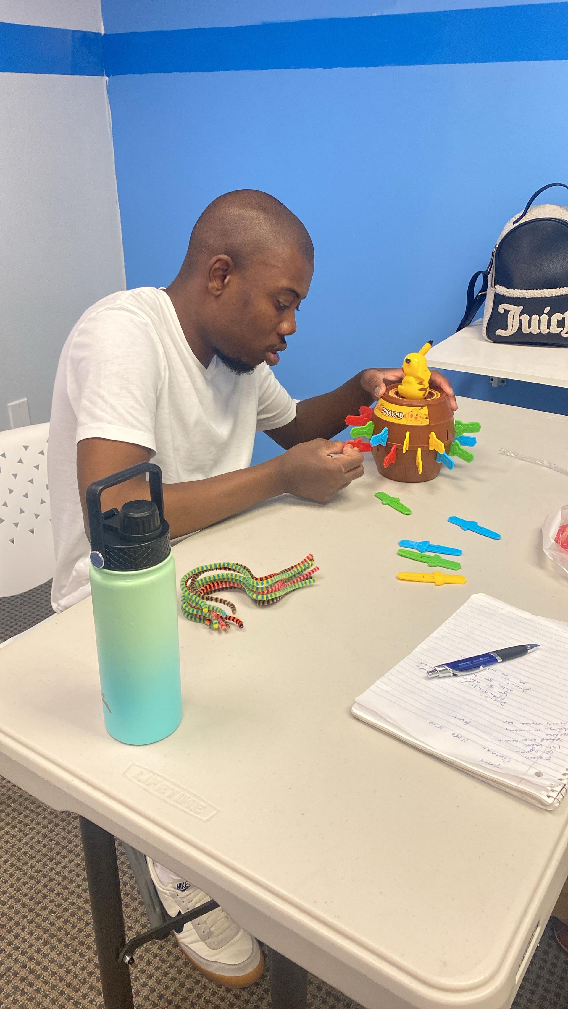 Man playing a stacking game at a table, with colorful pieces, water bottle and notebook visible.
