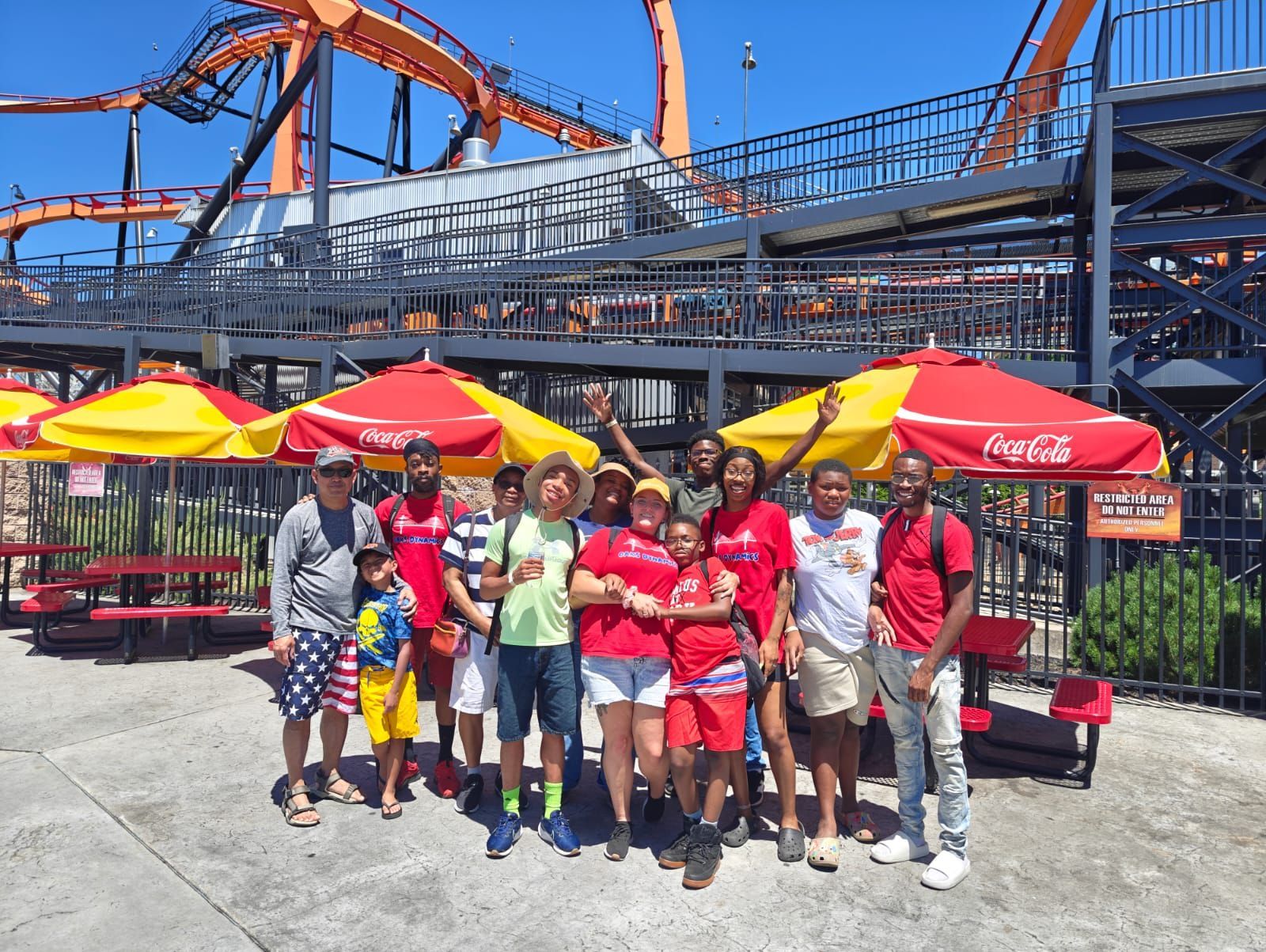 Group of people smiling, posing in front of a roller coaster, under red and yellow umbrellas.