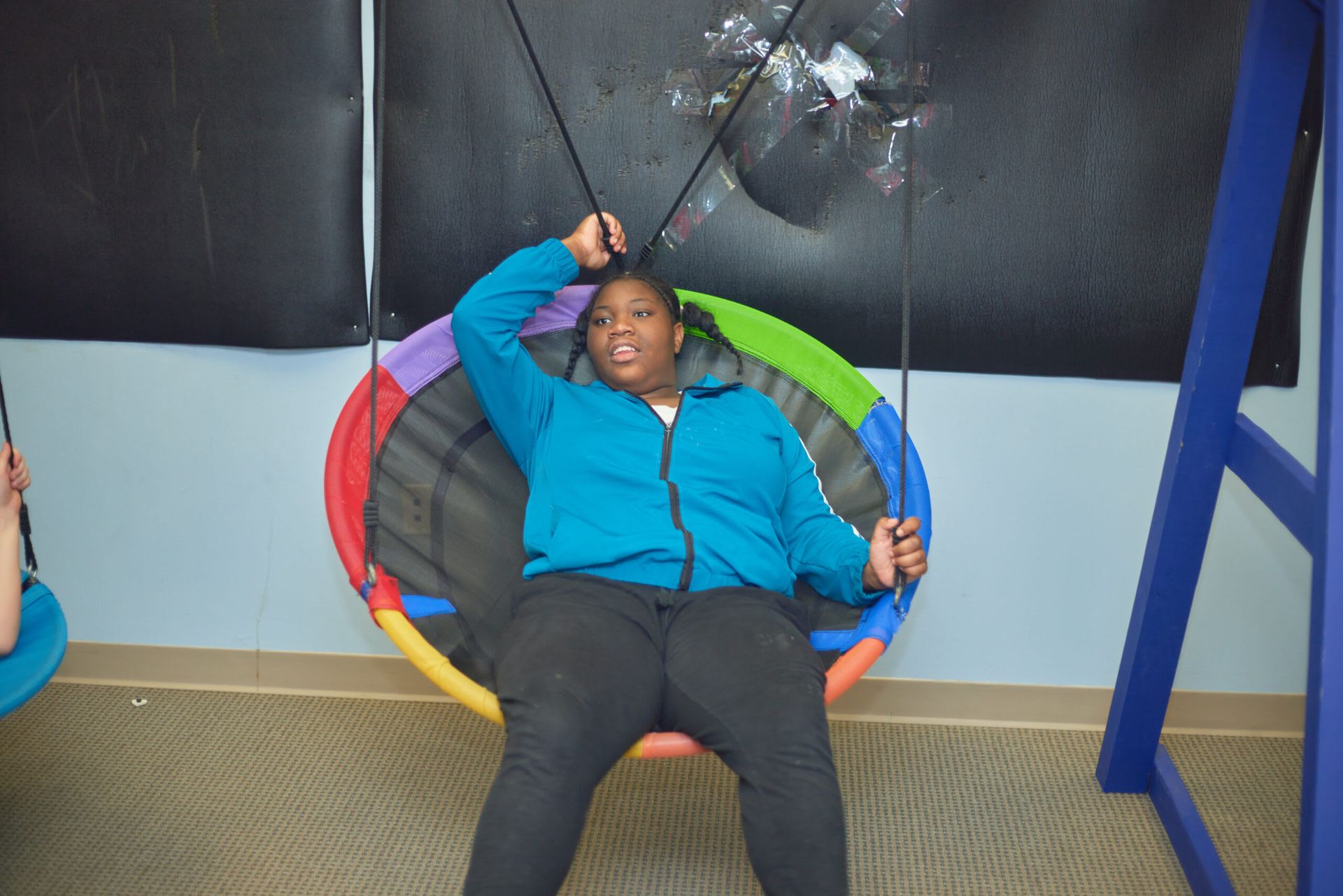 Person in a blue jacket sits on a colorful circular swing, arm raised, indoors.