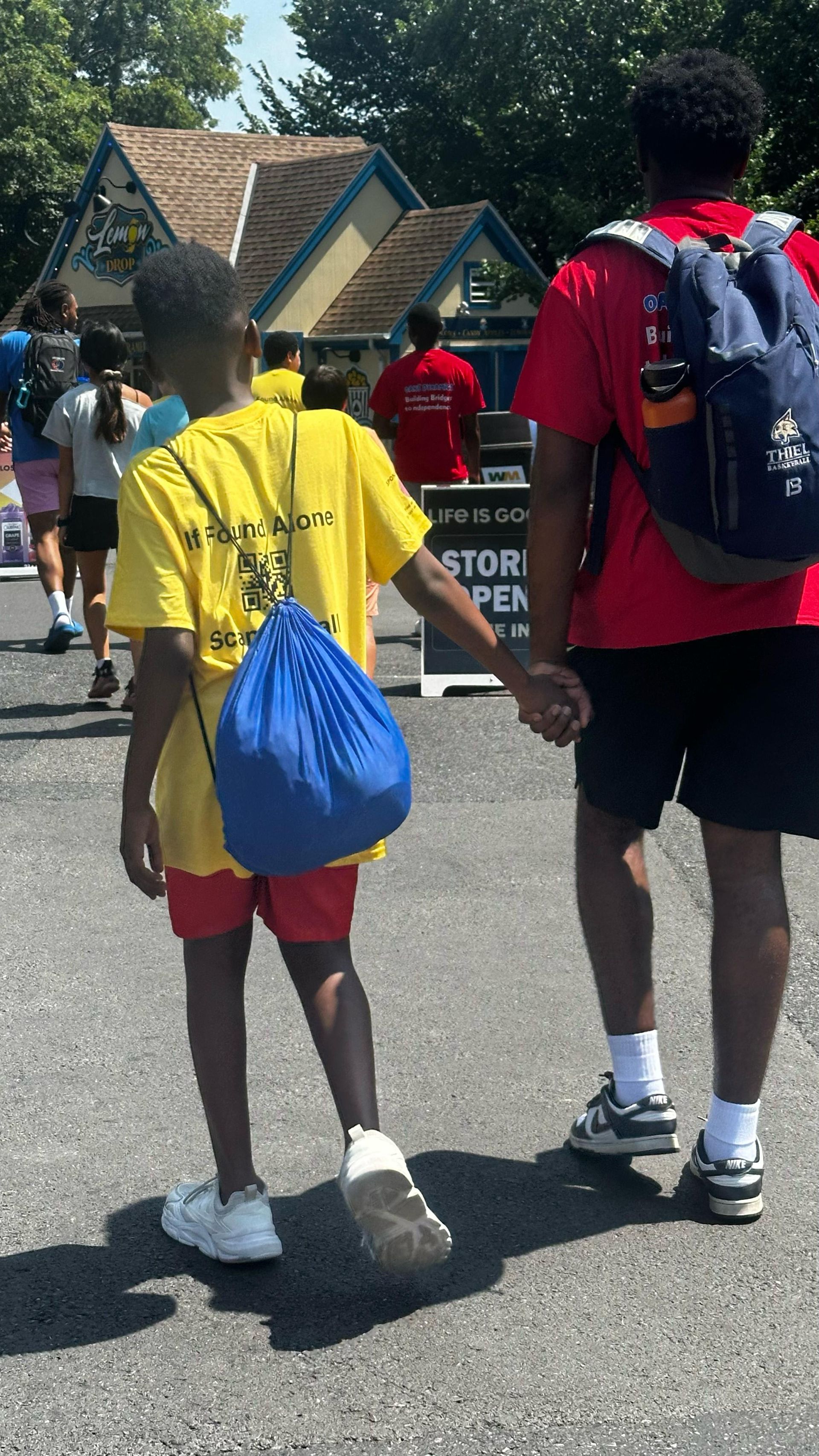 Two people, one in yellow shirt and blue bag, other in red, holding hands, walking on a path in front of a building.