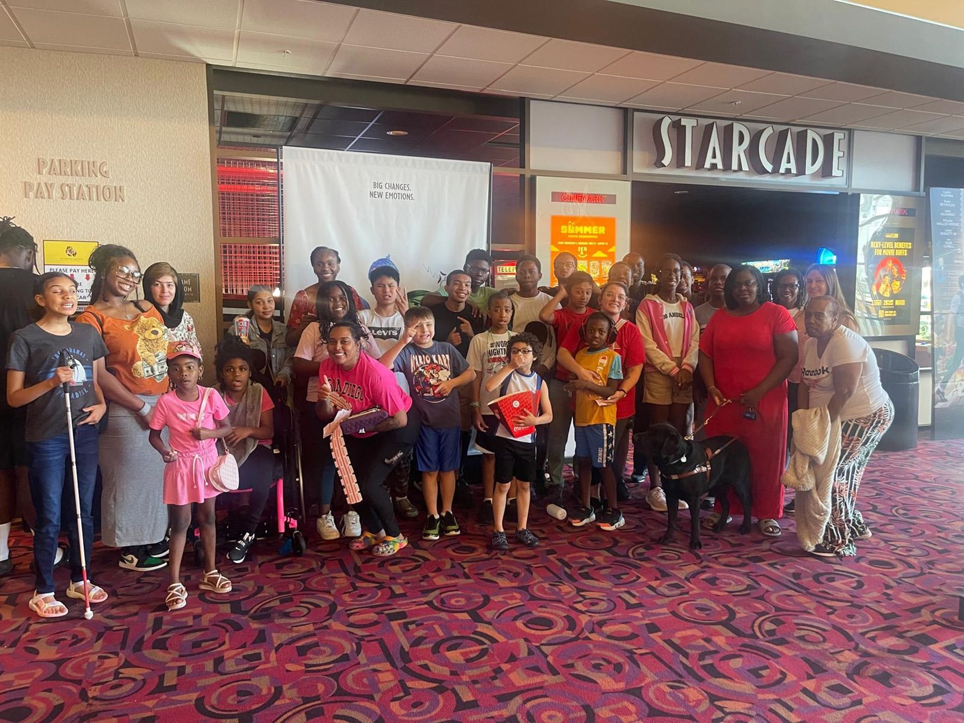 Group of children and adults posing in front of a movie theater. Red carpet, various skin tones.