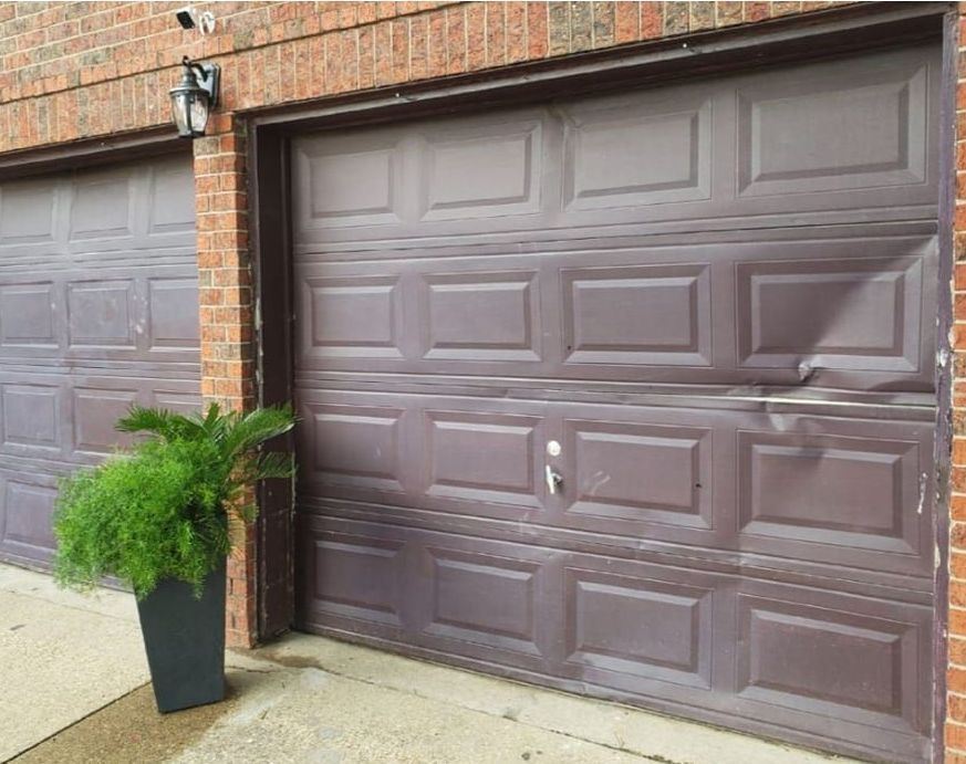 Brown garage doors beside a brick wall, with a potted fern on the driveway