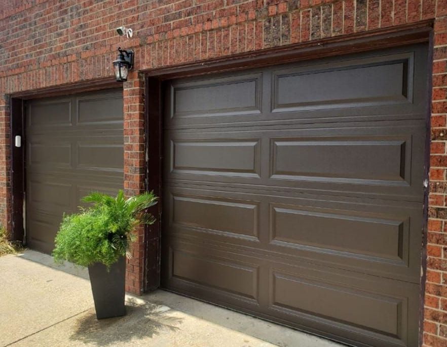 Two brown garage doors on a brick house, with a black planter of green fern on the driveway.