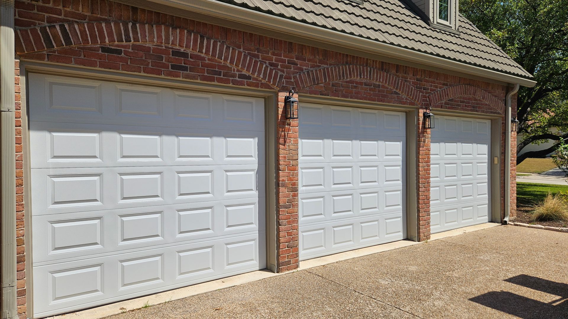 Three white garage doors on a brick house with a sloped roof and gravel driveway.