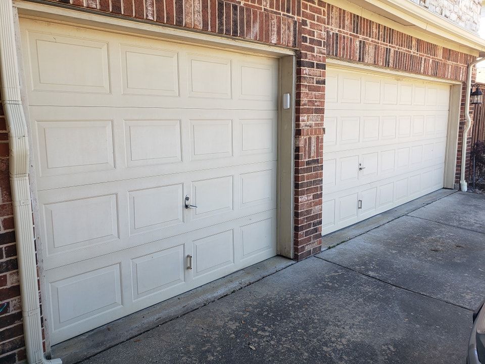 Two white garage doors beside a brick wall along a concrete driveway.