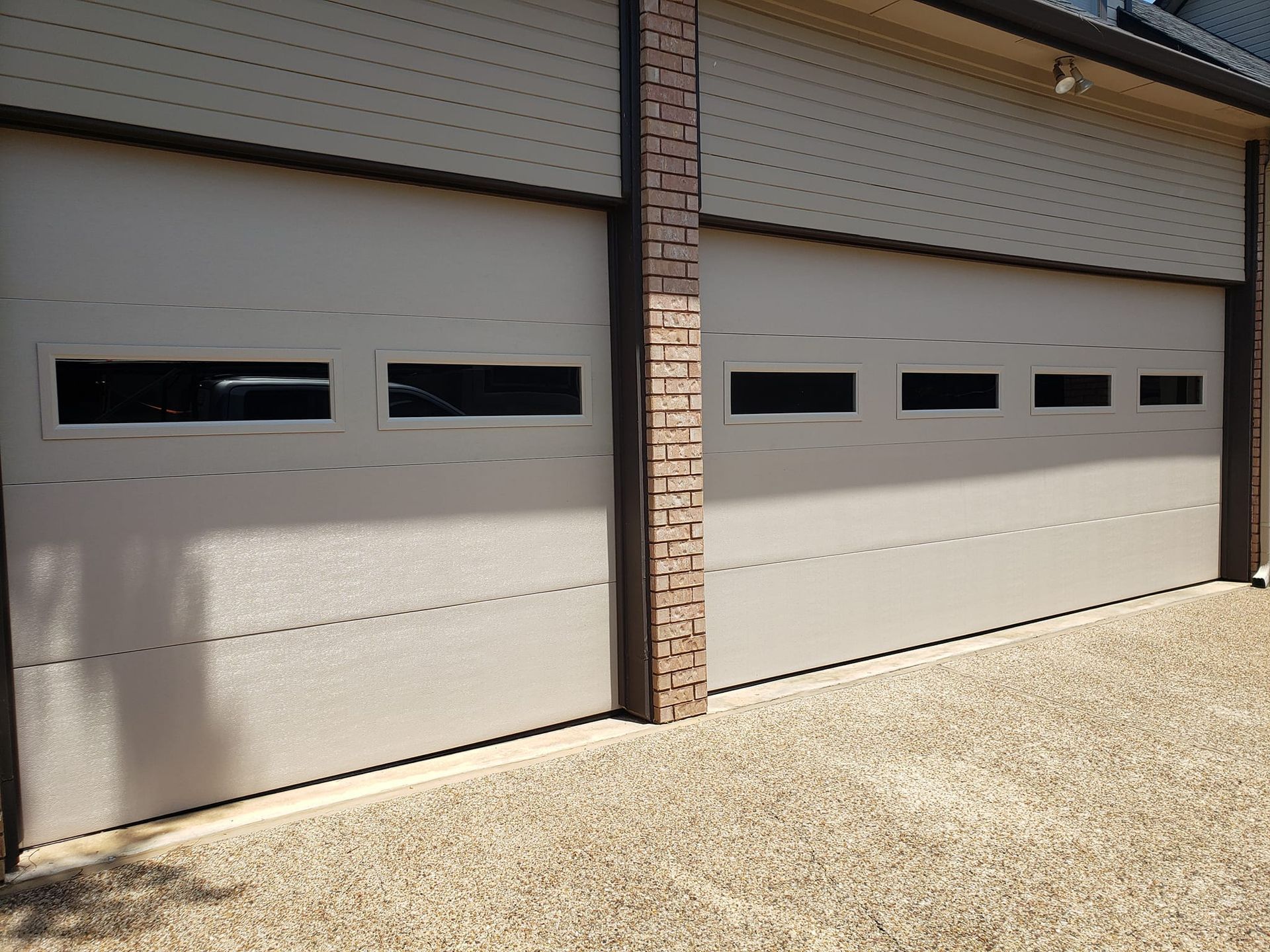 Two beige garage doors with narrow windows beside a brick pillar on a gravel driveway