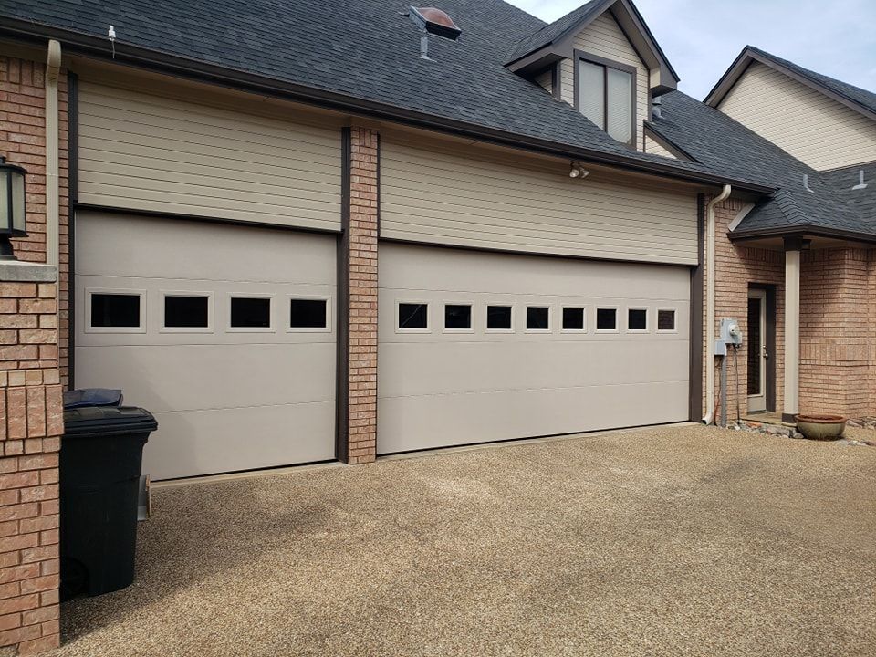 Two beige garage doors on a brick townhouse with a paved driveway and black trash bin