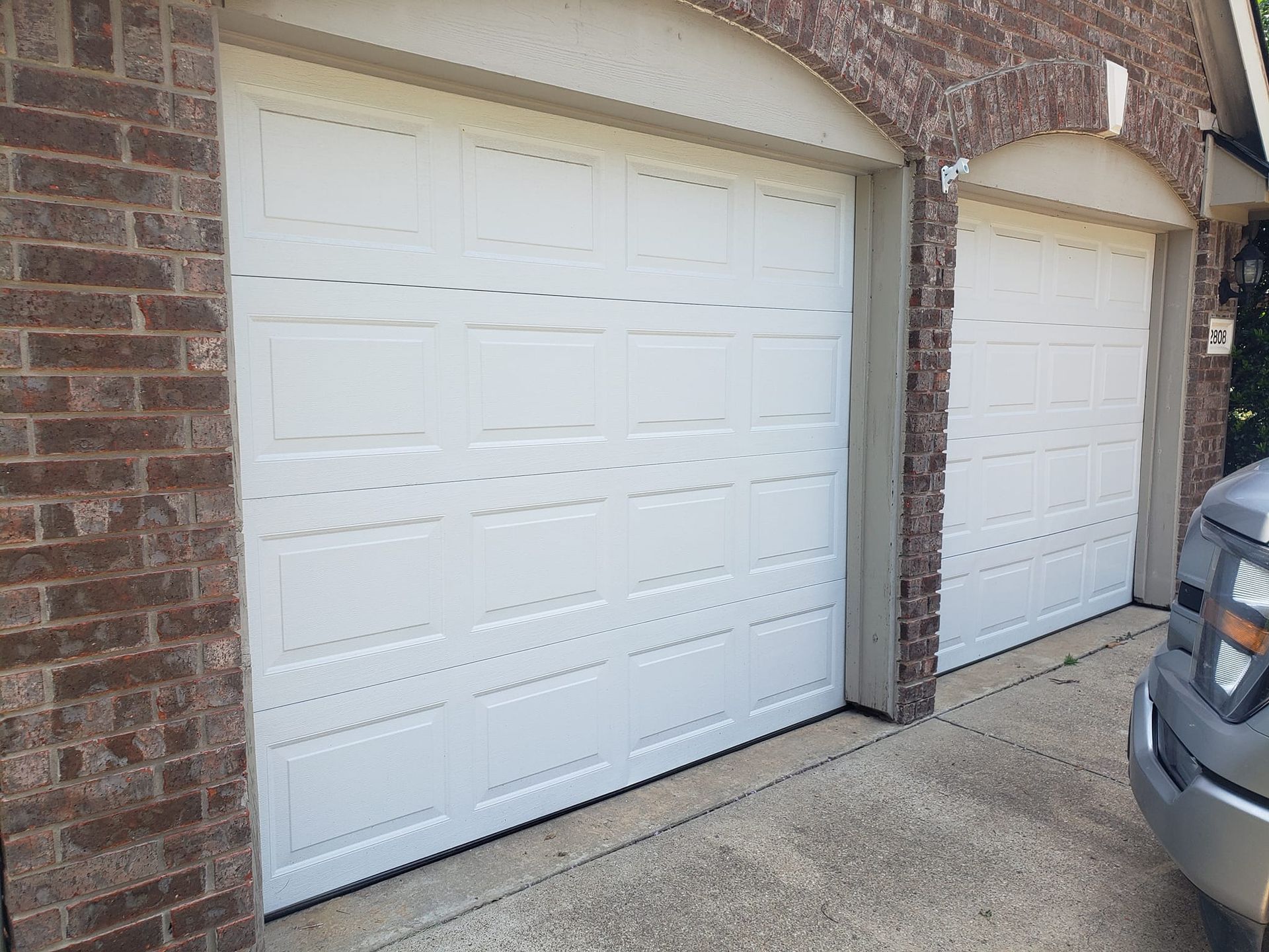 Two white garage doors on a brick house driveway, with part of a parked car visible on the right.