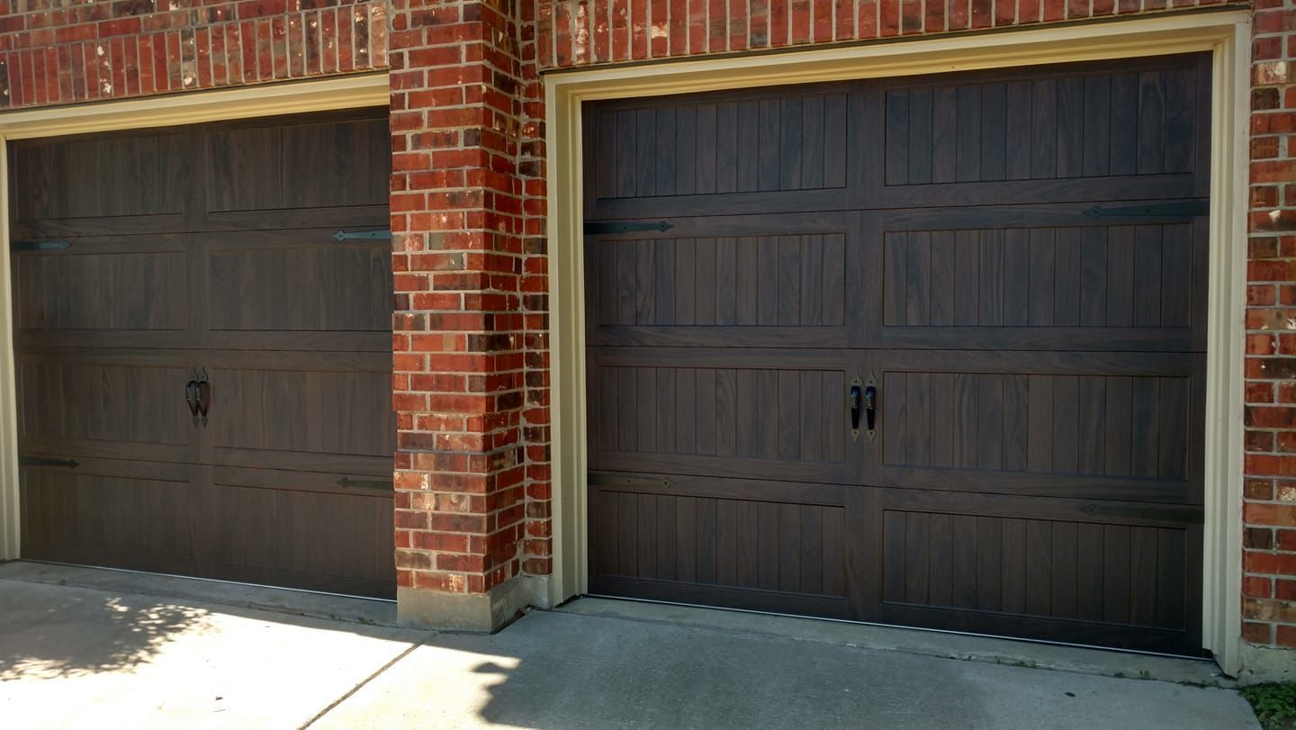 Two dark brown garage doors in a red brick building, with sunlight and a concrete driveway.