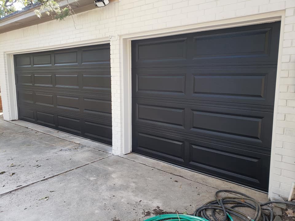 Two black garage doors on a light stucco house with a concrete driveway and coiled hose.
