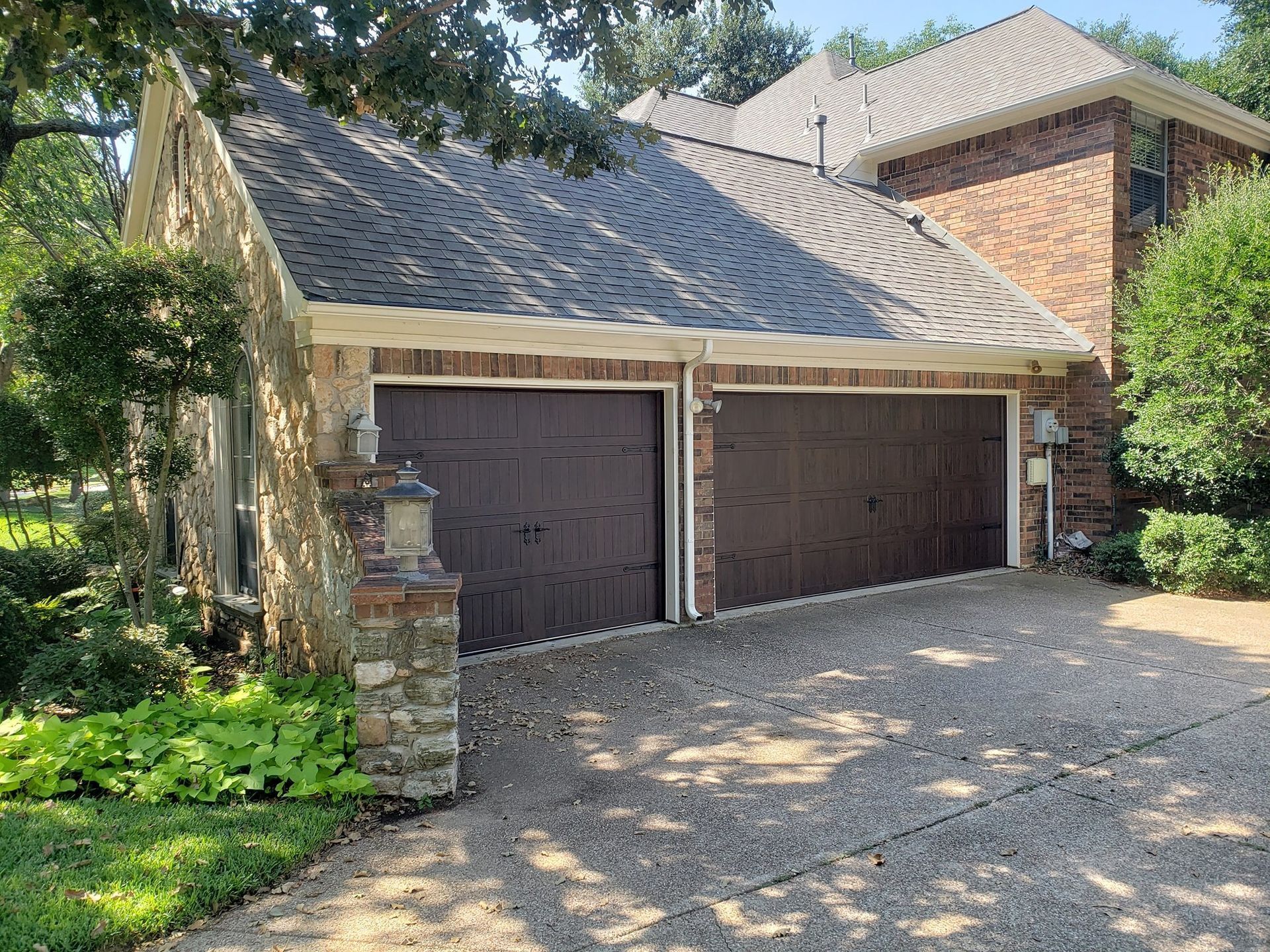 Two-car garage with dark wooden doors and stone columns, set beside a brick house and trees