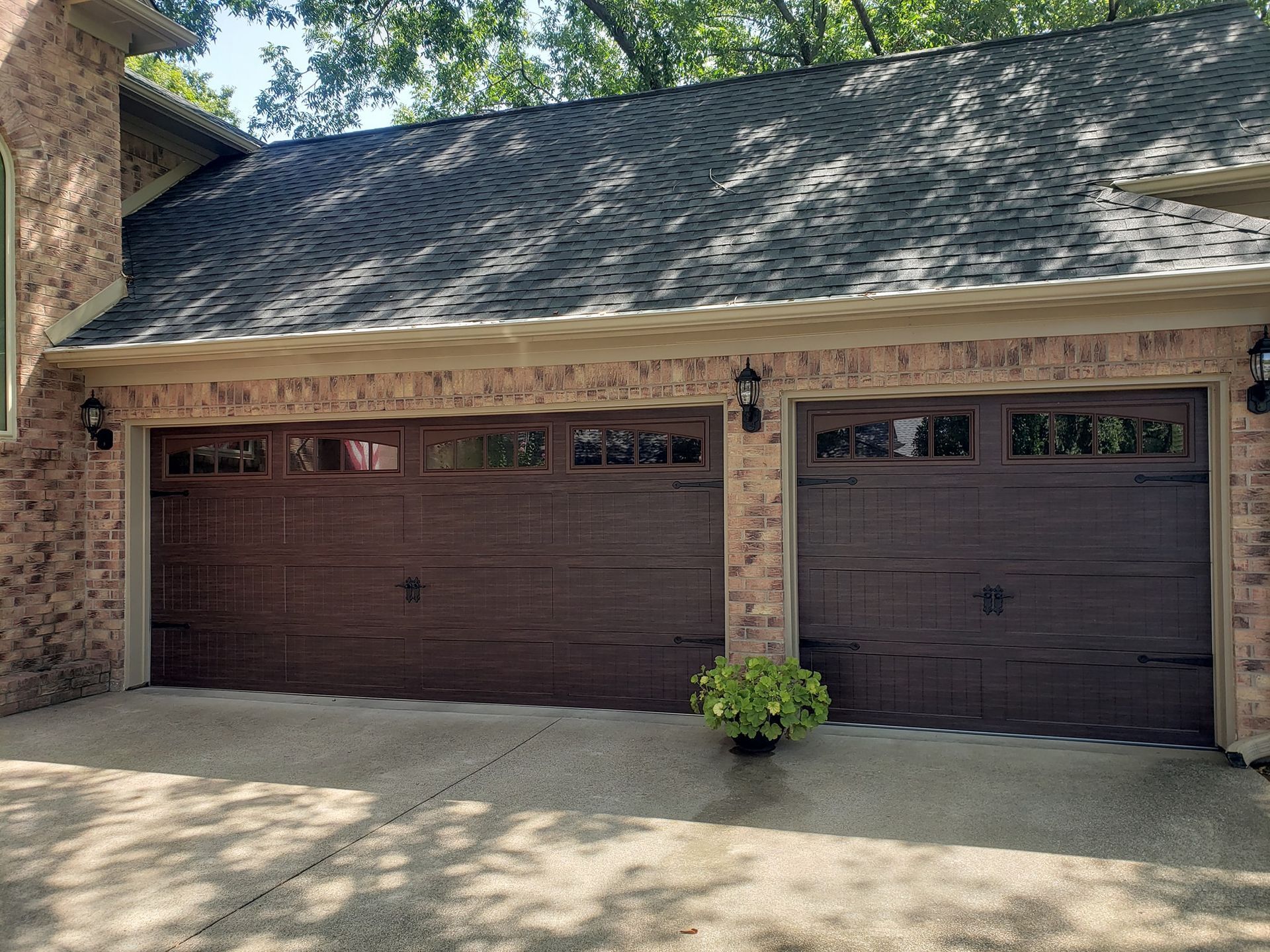 Two dark brown garage doors on a brick house with a shingled roof and small potted plant in front