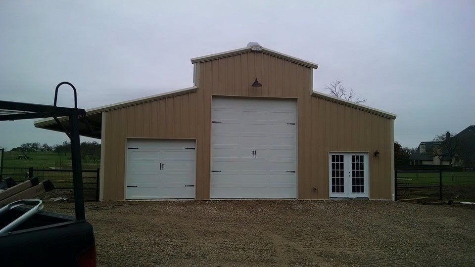 Tan metal garage building with white doors and a small side entry on a gravel lot under cloudy skies