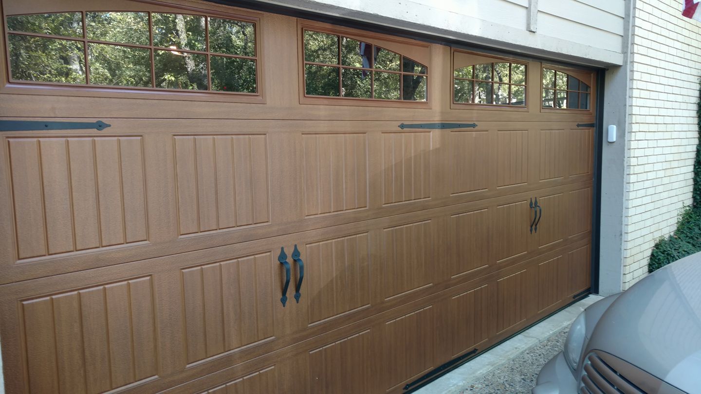 Brown wooden garage door with small windows beside a white brick wall and a parked car edge