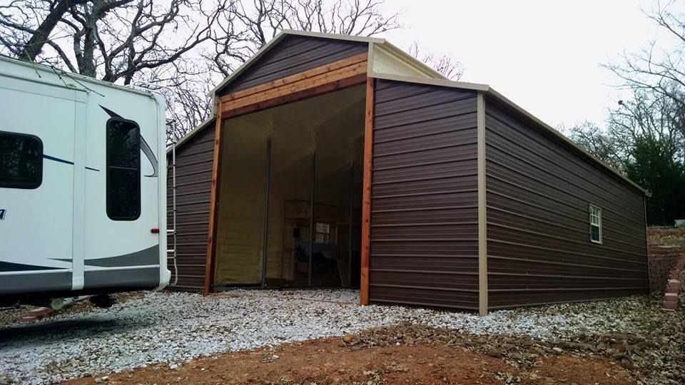 Large brown metal carport or garage beside an RV on a gravel lot under cloudy skies