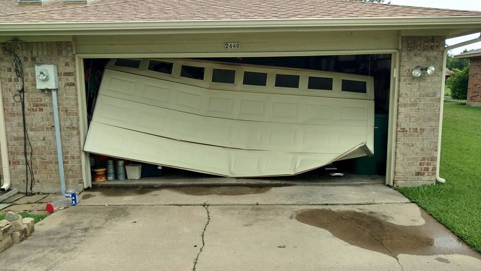 Bent garage door hanging open over cracked driveway, with a wet stain in front of a brick house.