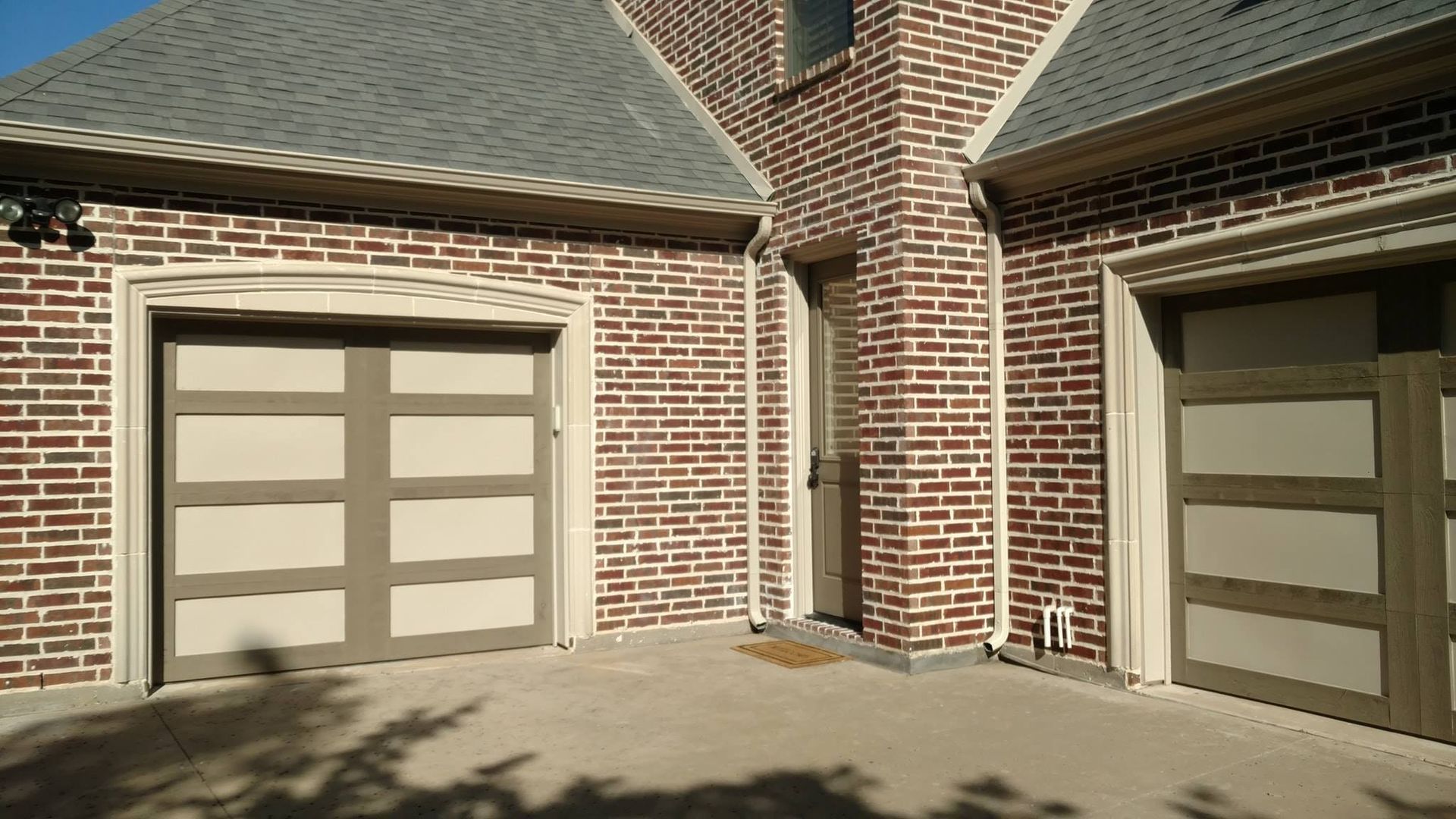 Brick house exterior with two beige garage doors under a shingled roof in sunlight