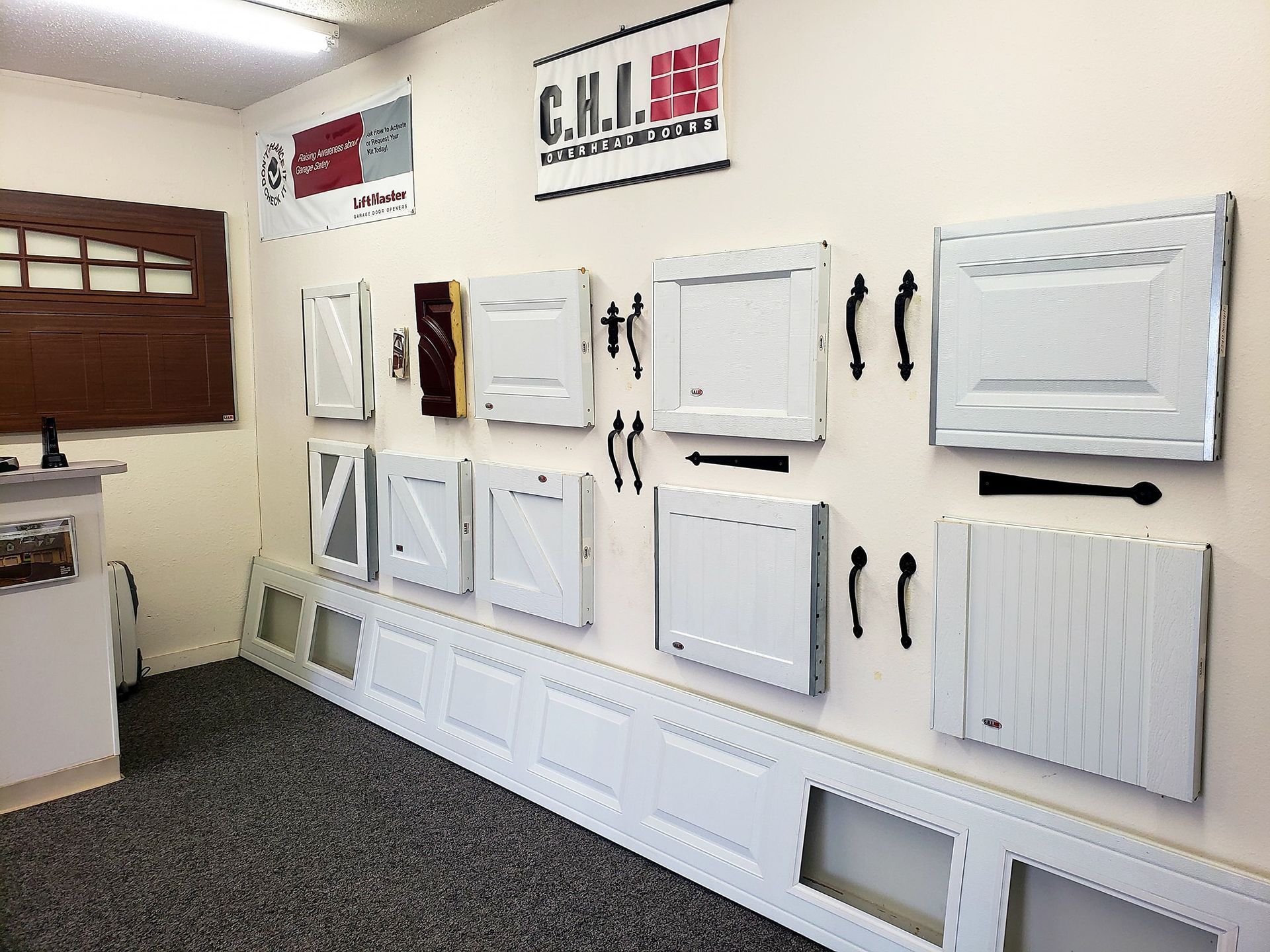 Display of white cabinet doors and drawers in a showroom, with sample panels on a wall and carpeted floor.