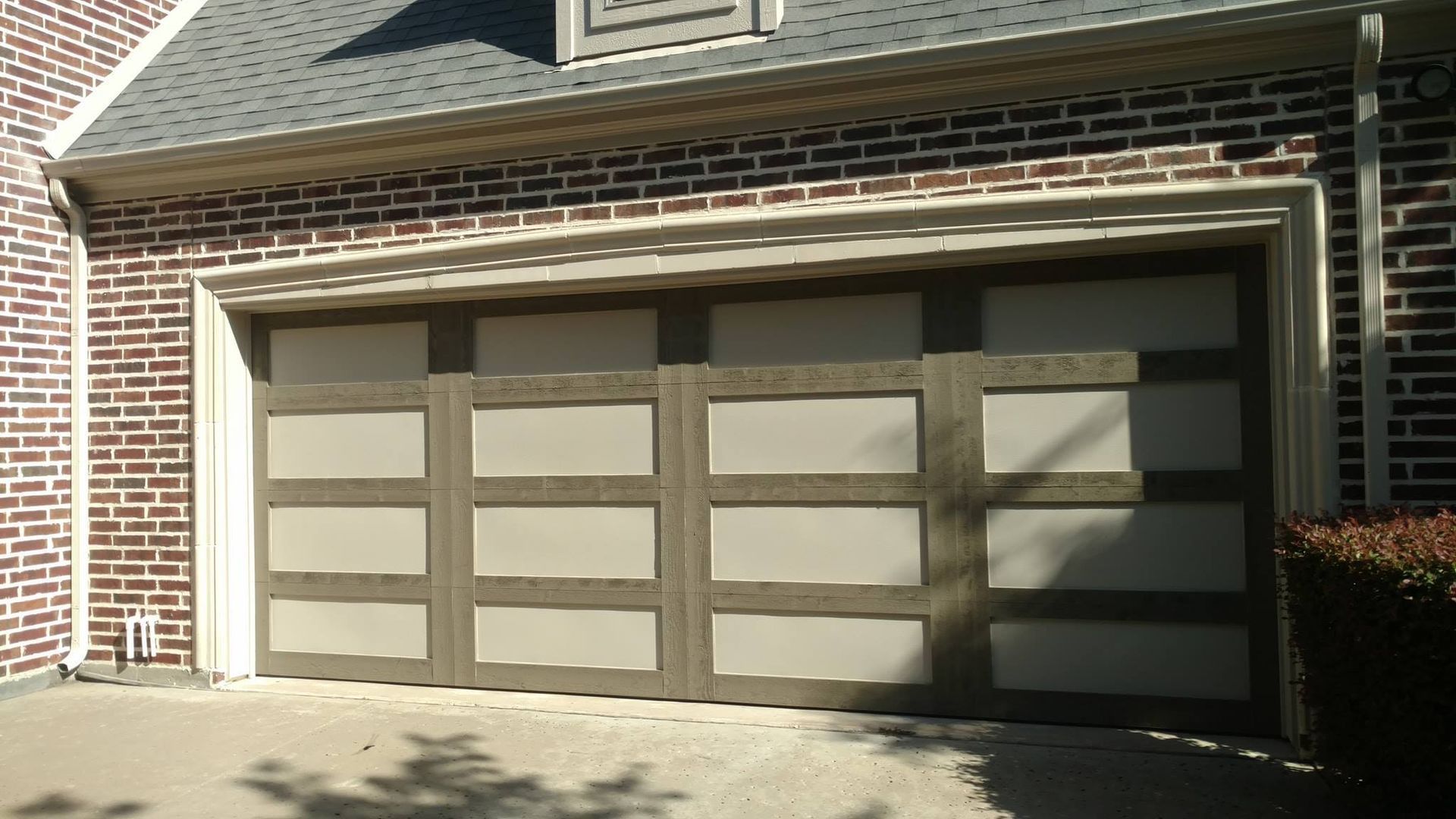 Brick house with a beige two-car garage door in sunlight and tree shadows on the driveway