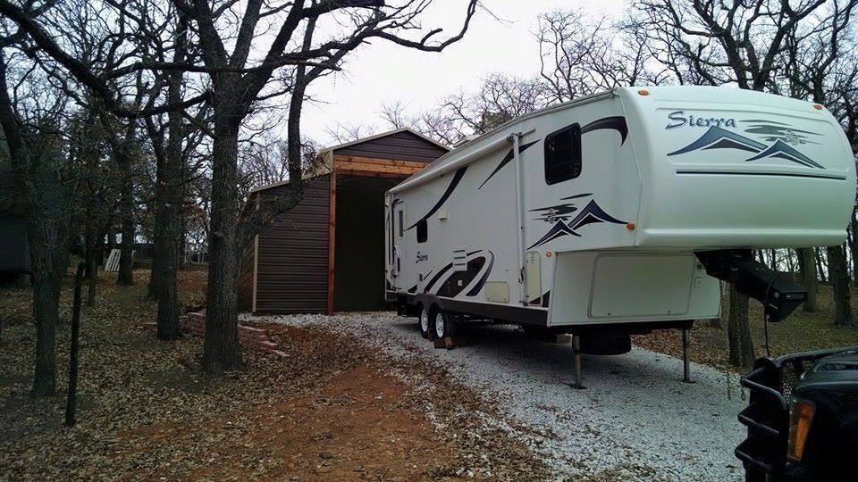 White fifth-wheel RV parked beside a wooden shed in a leafless wooded campsite.