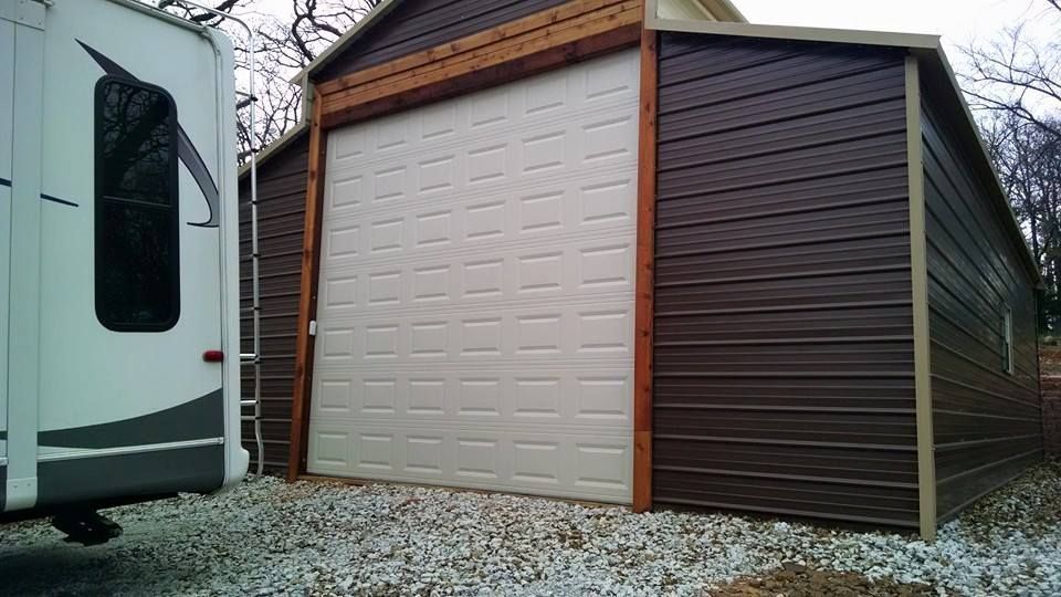 Two-sided storage shed with a white roll-up door and dark brown side door beside an RV on gravel