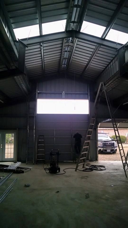 Inside a large warehouse, a person stands in silhouette near a bright open doorway and parked car.
