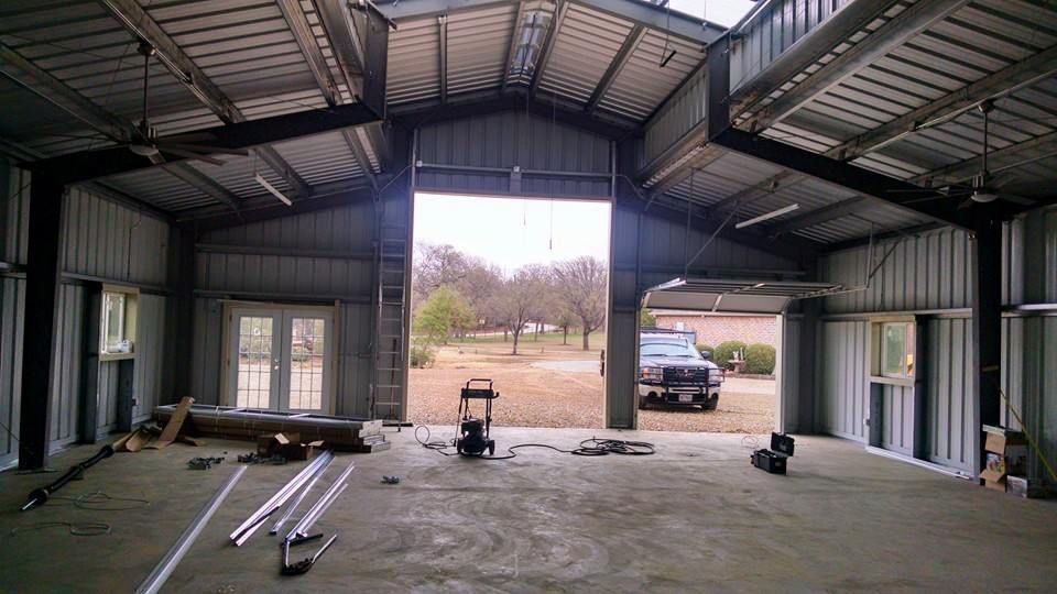 Empty metal workshop with open garage doors, construction materials on floor, and a truck visible outside.