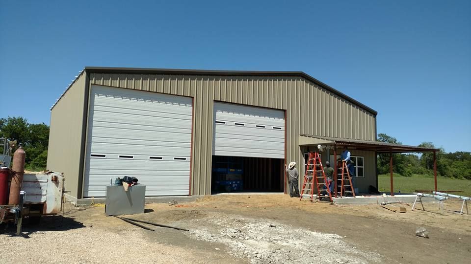 Tan metal warehouse with two large roll-up doors, one open, on a gravel lot under a blue sky.