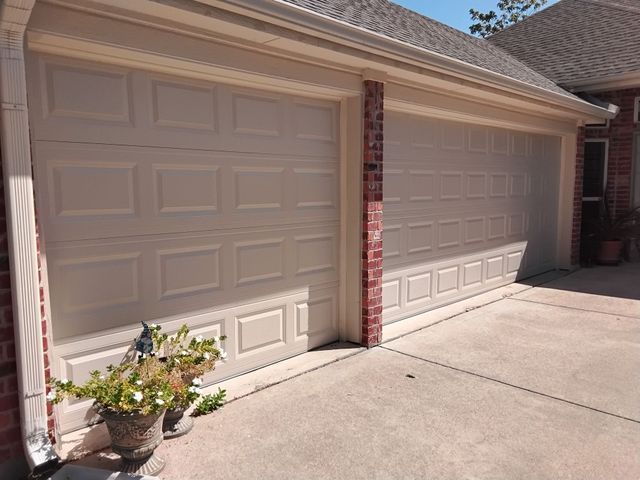 Two beige garage doors on a sunlit driveway beside a brick house, with a potted plant near the left door.