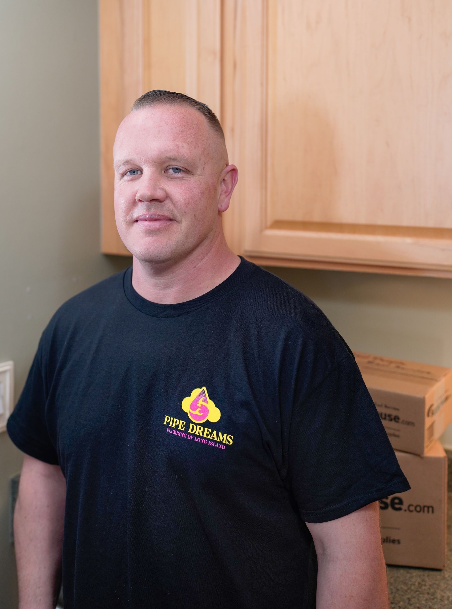Man standing in a kitchen beside wooden cabinets and cardboard boxes, wearing a black T-shirt.
