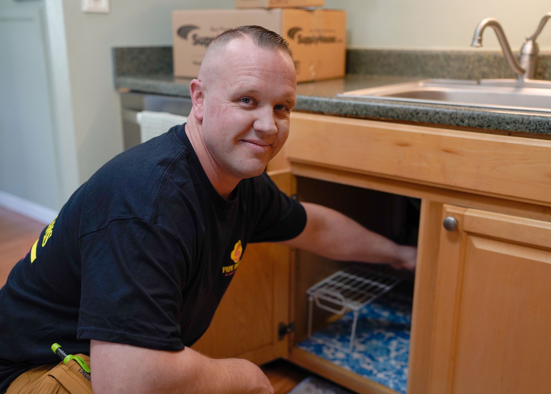 Man crouching under a kitchen sink, reaching into the cabinet while working on plumbing