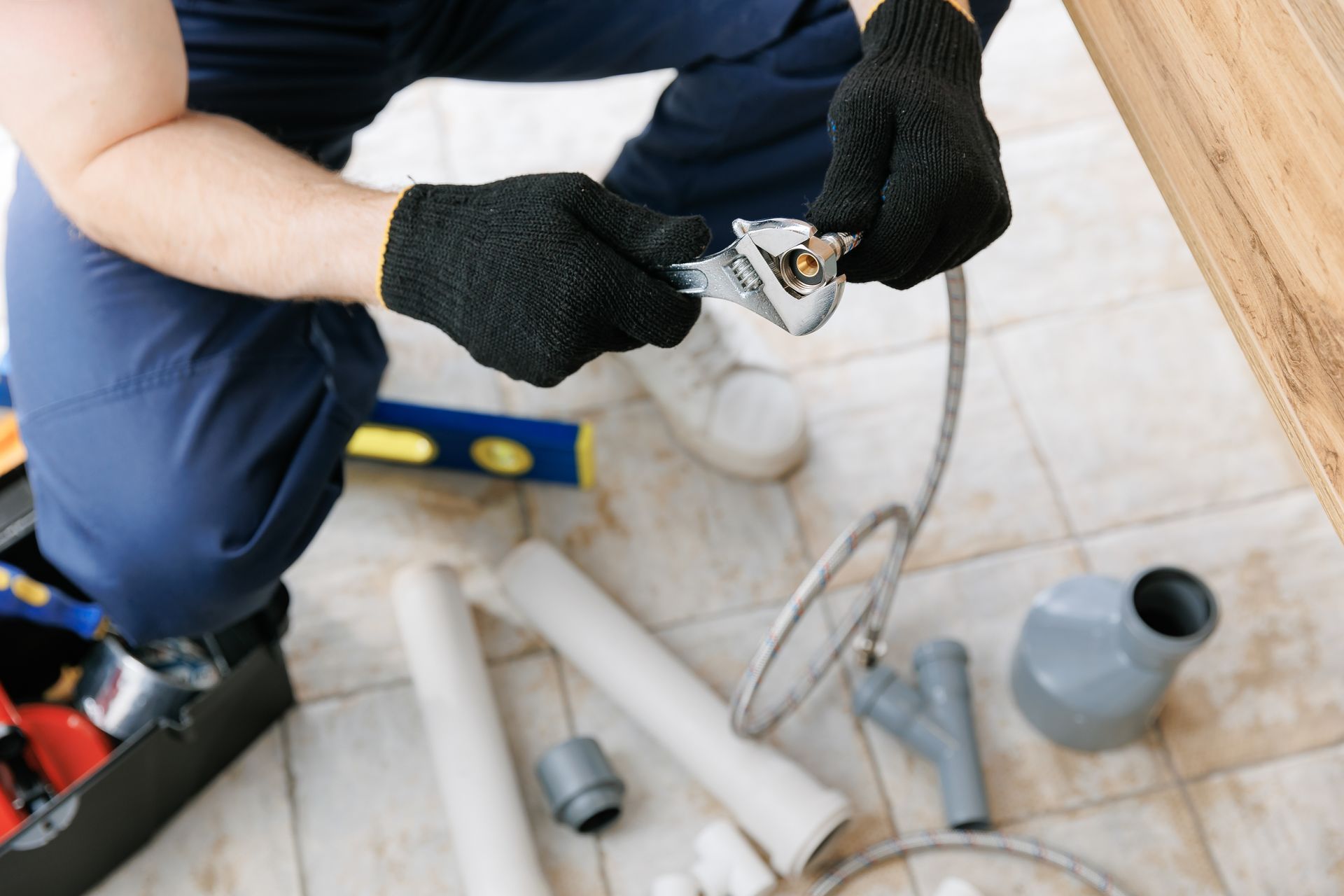 Gloved plumber installing a pipe under a sink with tools and fittings on the floor