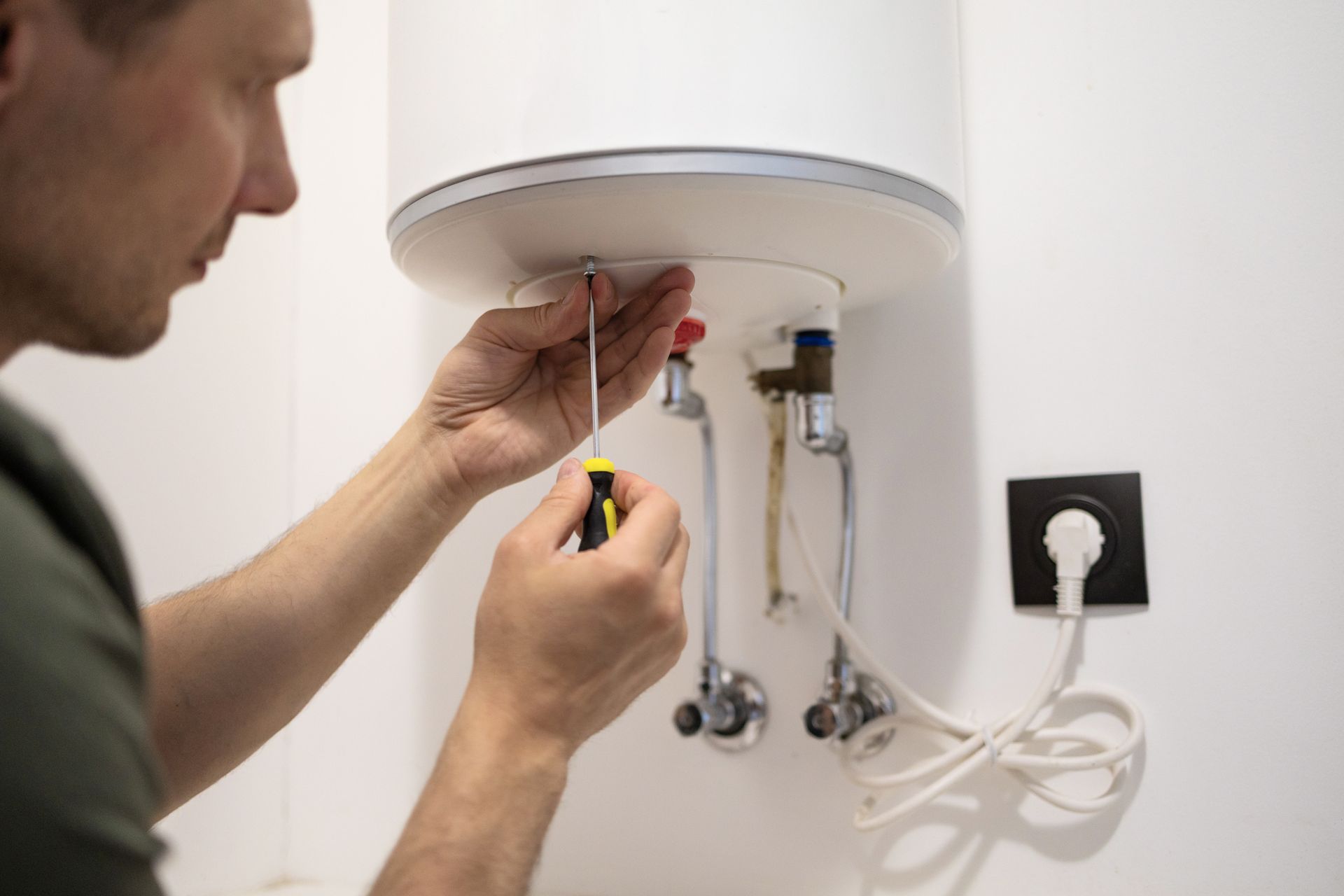 Person using a screwdriver to work on wires under a wall-mounted water heater near an outlet.