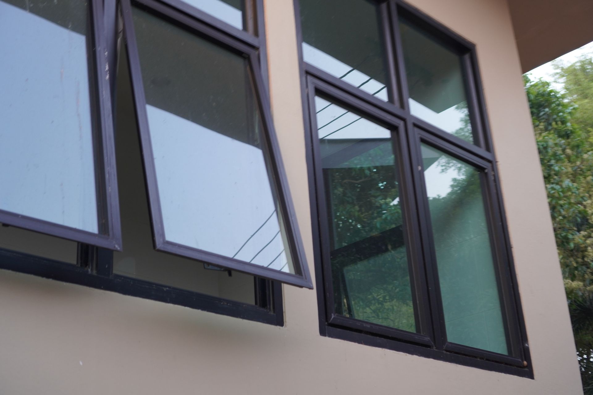 Black-framed windows, one open, set in a beige building exterior, reflecting sky and trees.