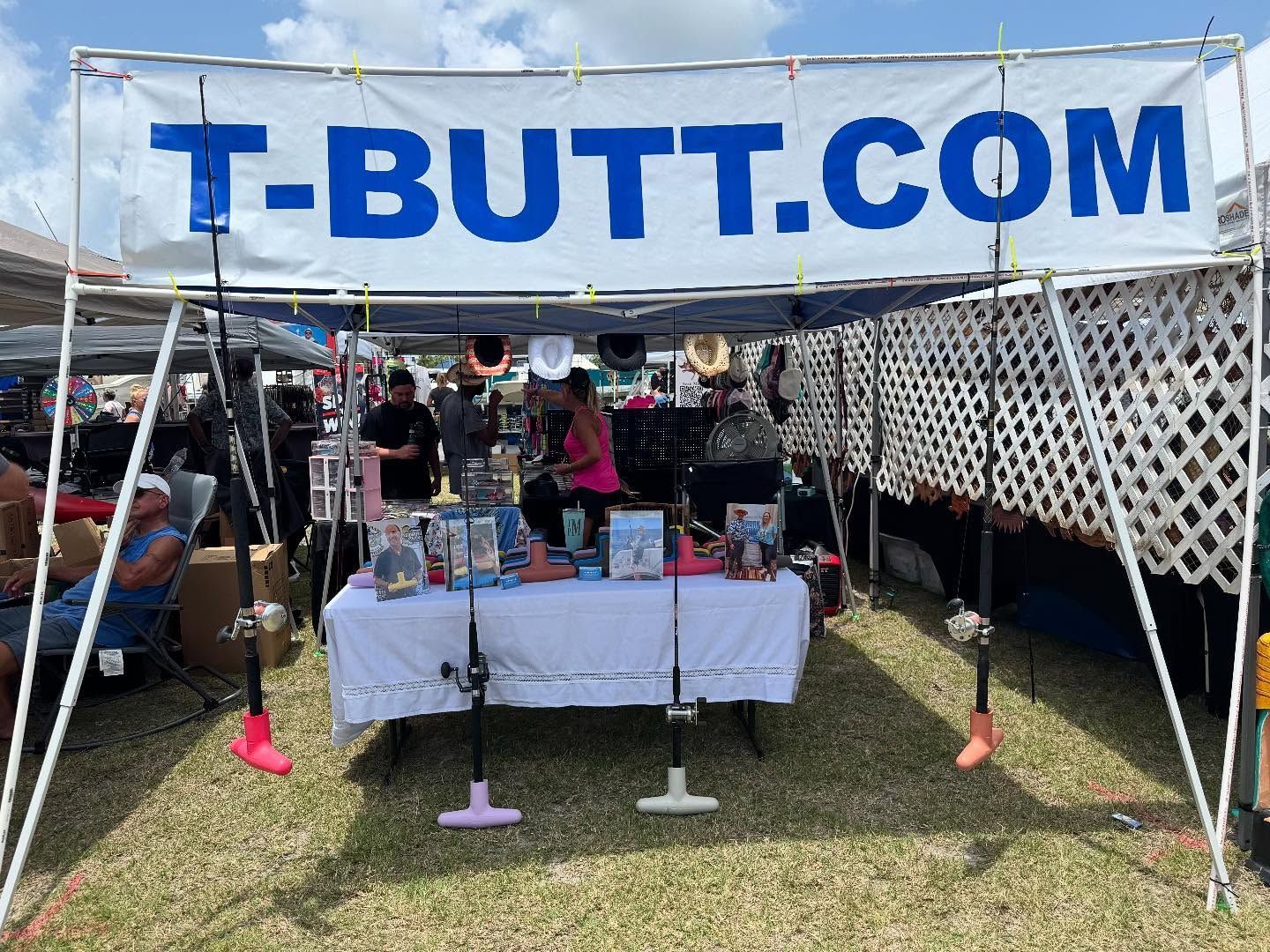 Vendor booth under a blue T-BUTT.COM banner at an outdoor market, with tables of items displayed.