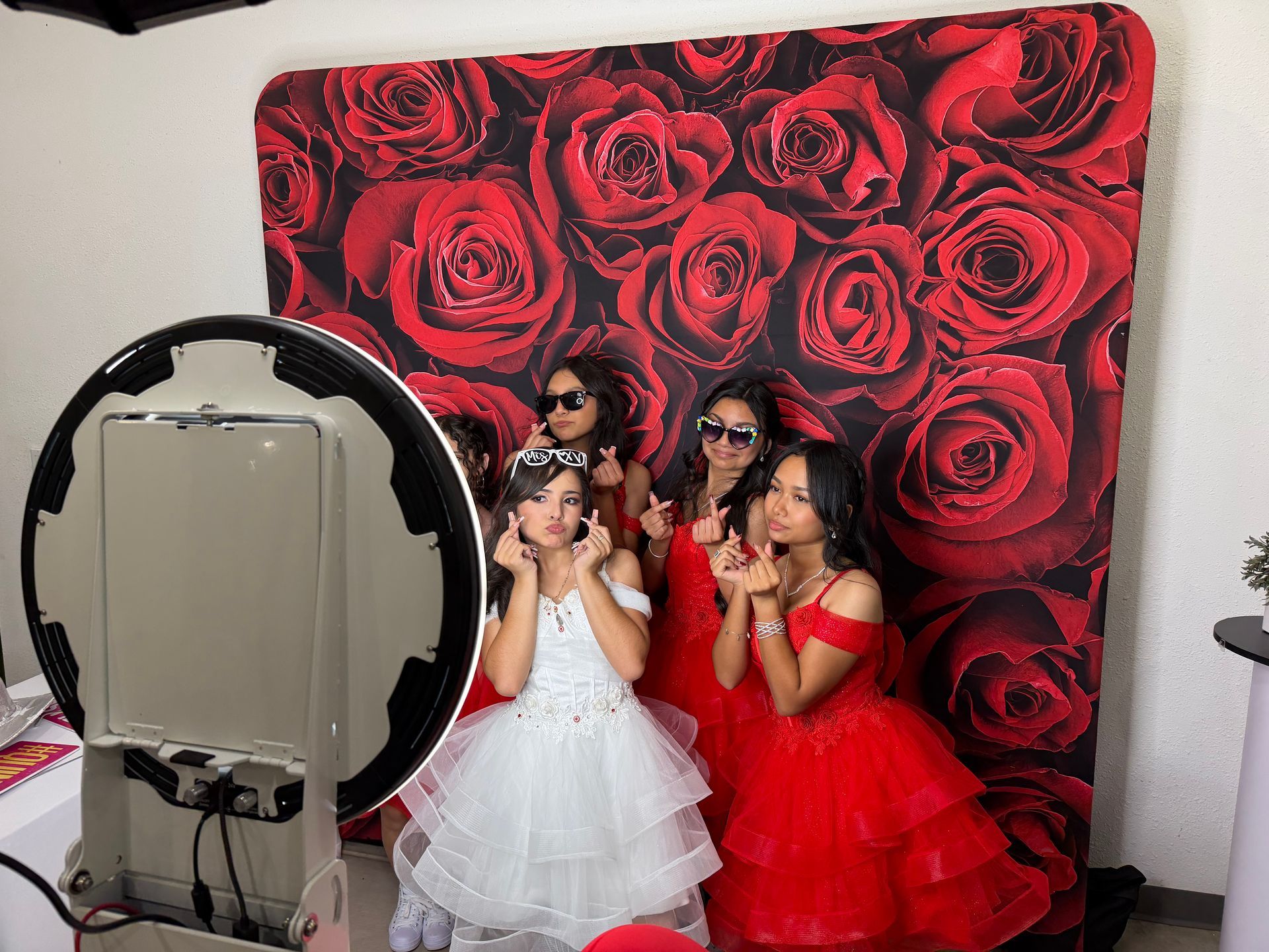 Ivana in her white dress posing with friends at her Quinceañera photo booth with a red rose backdrop in Perris, CA