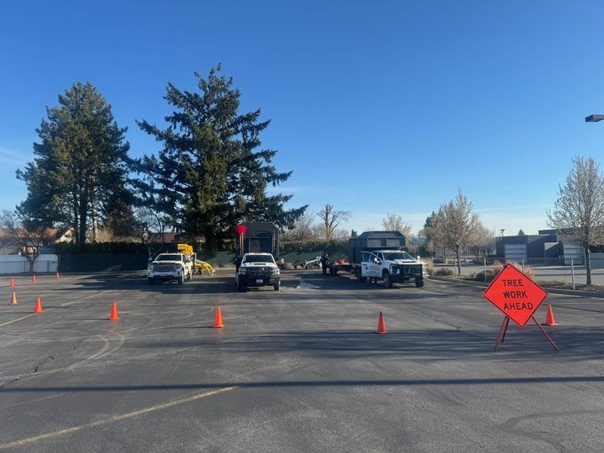 Orange traffic cones and a roadwork sign in a parking lot with trucks and trees under a clear blue sky
