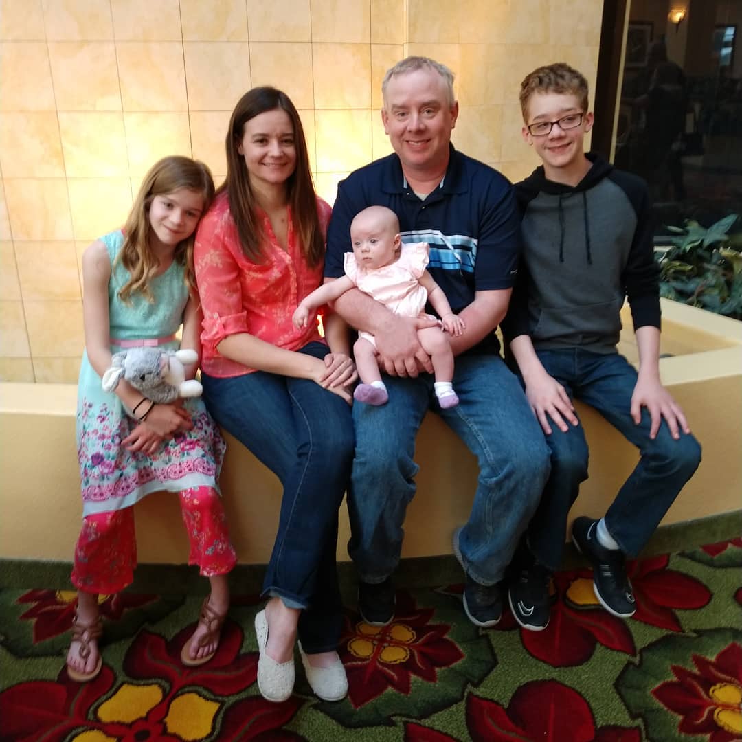Family of five sitting together, smiling. Girl in pink pants holds stuffed animal, baby held by dad.