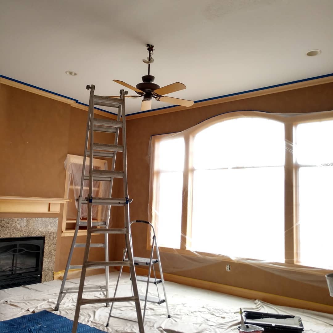 Interior room being painted, featuring ladder, covered windows, and taping around trim.