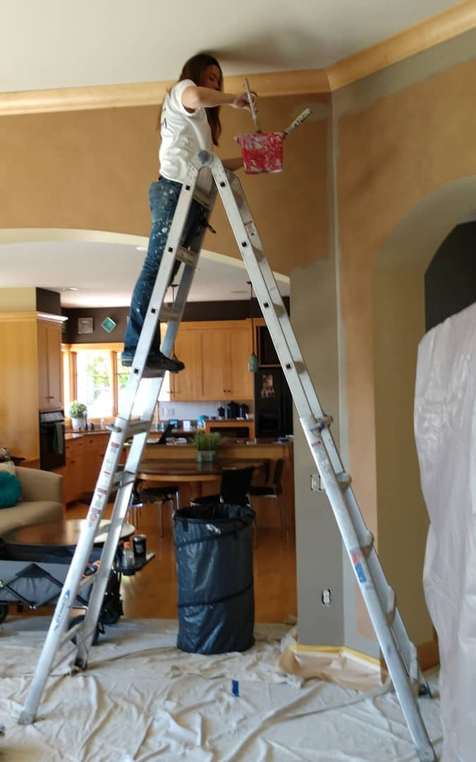 Person on a ladder painting a wall, interior setting with protective drop cloths on the floor.