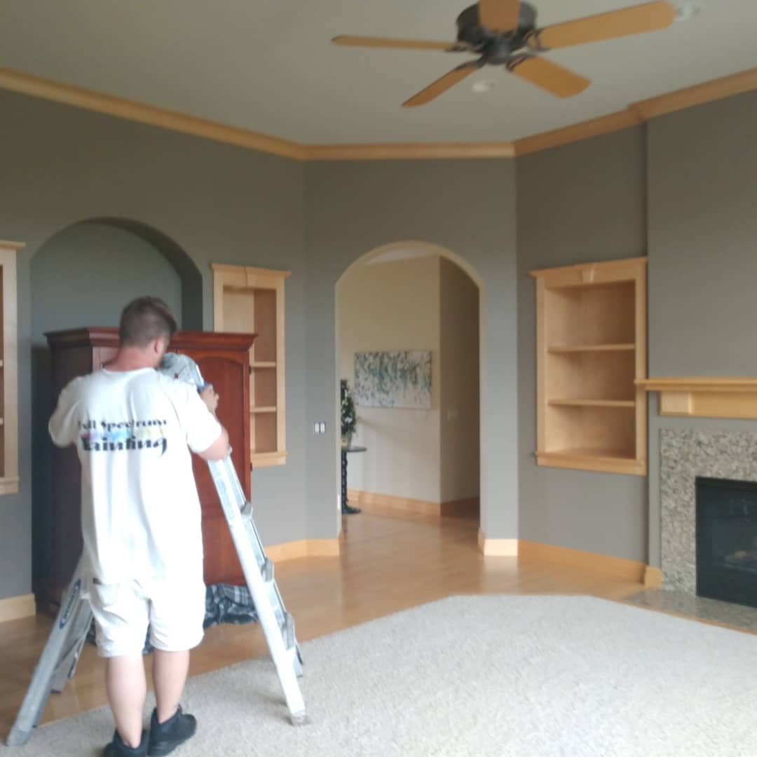 Person painting interior room with gray walls, wood trim, and a ceiling fan.